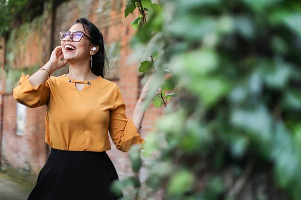 Young Latin American woman wearing eyeglasses and earbuds looking up smiling with leaves copy space. High quality photo