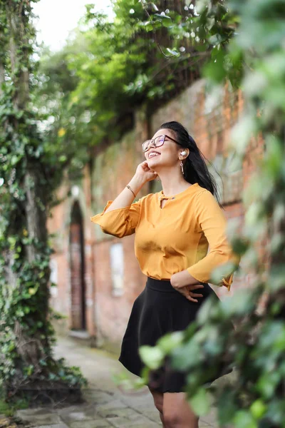 Young Latin American woman wearing eyeglasses and earbuds looking up smiling with leaves copy space. High quality photo