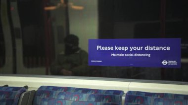 Person wearing face mask on public transport, with Covid-19 Coronavirus information sign in London Underground tube train carriage for social distancing in England, UK