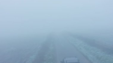Aerial drone video of car driving in misty thick foggy, bad and dangerous driving conditions on a rural countryside road, 4 wheel drive with trailer driving past fields in England in mist and fog