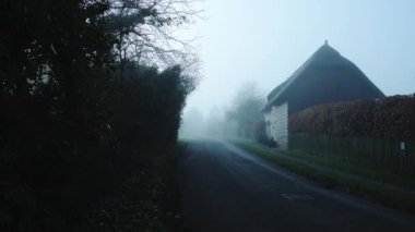 Rural narrow countryside road in thick fog and mist through a small village, dangerous bad driving conditions in foggy misty weather on English roads, back lanes streets in England, UK