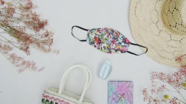Panning through a minimalist view of modern essentials: face mask for coronavirus pandemic, a pastel-colored purse, with wide-brimmed sun hat against a pure white background and dried flowers.