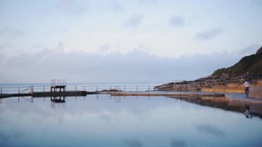 A Man Walking To The Deck Of The Swimming Pool To Capture The Beauty Of Guernsey Waters. -wide shot