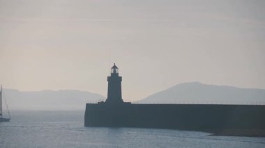 Silhouette Of Guernsey Lighthouse With Two Sailboat Cruising In St. Peter Port, Channel Islands, Guernsey During Sunrise.- wide shot