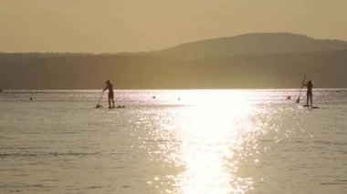 Silhouette Of Couple Doing Stand-up Paddle Board Surfing On The Calm Ocean In Italy With Beautiful Reflections Of Bright Summer Sunset. - wide shot