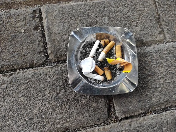 An aluminum plate ashtray, full of trash and cigarette butts, was on top of the concrete paving.