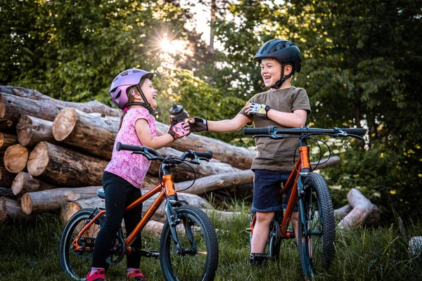 Two smiling children with bike helmets pass the bottle while cycling in the forest