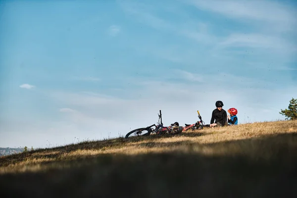 Baba ve oğul çayırda oturuyor ve dağ bisikletlerinin yanında konuşuyorlar.
