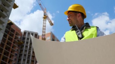 Handsome young male constructor engineer wearing helmet and uniform holding in hands work project and looking at construction site. Bottom view. Architecture business, inspector work, close up