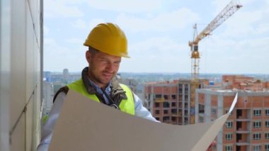 Close up of Caucasian handsome young professional male constructor in helmet looking at plan draft at high building side. Man builder with documents working at constructing. Work, construction concept