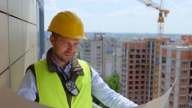 Handsome Caucasian happy male worker engineer working on project documentation at a construction site. Young man builder in helmet looking at arcitect plan draft outdoor. Close up. Building industry