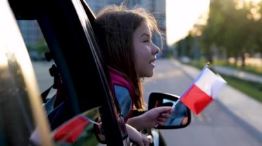 Pretty little girl with small flag of Poland on stick sits in front seat in automobile. Happy female teen holding Polish flag looking out of car window. Close up concept. Kid patriot. Traveling around