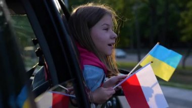 Teenager sits in car looking out the window waving Ukrainian and Polish flags. Little Caucasian girl holding in hands flag of Ukraine and Poland. Kid refugee flee from war. Close up, slow motion