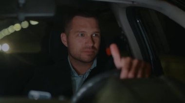 Close up of handsome young Caucasian businessman in suit driving car at night and smiling looking around. Man traveling in car from work. Taxi driver works in evening. Slow motion. Luxury style