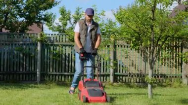 Handsome male gardener mowing grass with an electric lawn mower. Young man mows the grass in own garden with a grass collector on a sunny summer day. Beautiful landscape design. Slow motion. Lawn care