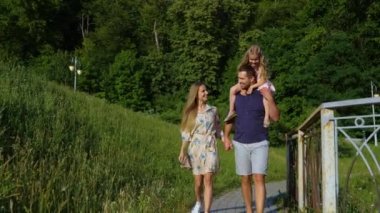 Cute family couple with little child walking in park on sunny day. Happy family holding hands on a walk with cute small daughter. Summer weekend. Parenthood concept. Dad with kid on his shoulders