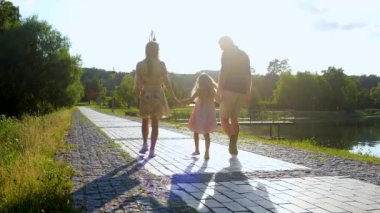 Happy family spending time together on sunny day. Mom, dad and daughter walk in park holding hands. Happy childhood concept. Family walking near beautiful lake. Back view. Slow motion. Parenthood