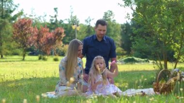 Caucasian young man and woman sitting on a blanket with child in green park and playing with soap bubbles. Happy cute family having picnic outdoor on summer. Family day. Childhood and parenthood