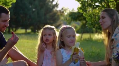 Close up of cute blonde little girls on family picnic with parents in park in summer. Caucasian children are eating fruits sitting on blanket outdoor. Happy childhood. Family weekend. Slow motion