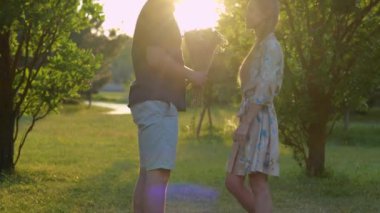 Handsome young man giving beautiful flowers to his pretty happy woman standing outdoor in park on sunset. Caucasian couple in love on nature. Romantic relations concept. Boyfriend and girlfriend