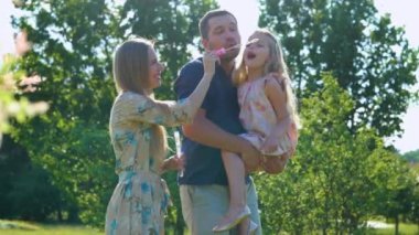 Joyful family spending summer day together playing with soap bubbles outdoors. Happy parents play with child on a walk. Cute pretty girl blowing bubbles with father and mother in park. Happy childhood