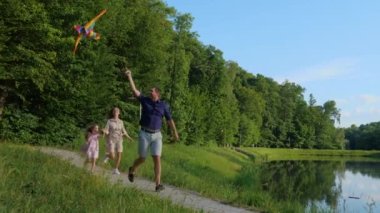 Joyful parents with girl kid having fun outdoors. Young mom and dad running with little child flying kite on nature. Parents with daughter playing with kite in park with beautiful lake. Family time