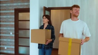 Portrait of Caucasian cheerful young couple in good mood entering their new apartment holding boxes in hands and putting them down on the floor. Moving in together, relocation. Family home. Marriage