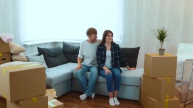 Camera approaching Caucasian happy married couple sitting on sofa in room with many boxes. Husband made a surprise for his beloved wife covered her eyes and showing their own apartment. Love concept