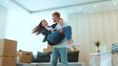 Young joyful married couple feeling happy together in new apartment. Low angle view. Loving young husband spinning his beloved beautiful wife on hands in new house on moving day. Own place to live