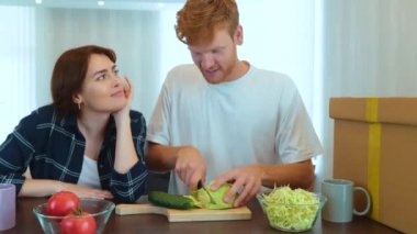 Happy young Caucasian couple standing in new house preparing food together. Cheerful family couple in new apartment. Husband cutting vegetables and eating cabbage in new kitchen. Slow motion