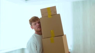 Close up of handsome Caucasian red-haired guy holding many cardboard boxes in hands indoors. Boxes falling on the floor. Relocation concept. Moving out the apartment. Change of home. Slow motion.