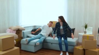 Young happy couple handsome man and pretty woman jumping on a sofa in living room with many cardboard boxes in it. Relocation concept. Moving in together. Slow motion. New house. Family concept