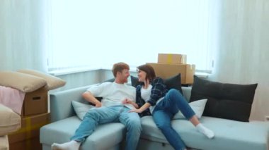 Camera approaching Caucasian joyful male and pretty female smiling jumping on a couch in light living room with many cardboard boxes. Boyfriend and girlfriend moving in together. Slow motion. Close up