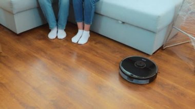 High angle view. Modern robot cleaning new apartment after moving in. Wireless robot vacuuming in room. Male and female legs sitting on sofa while vacuum cleaner working. Slow motion. Housekeeping