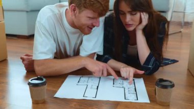 Cute young happy boyfriend and girlfriend lying on the floor in new apartment with sofa and many boxes looking at plan documentation discussing interior design of new home. Relocation. Moving day