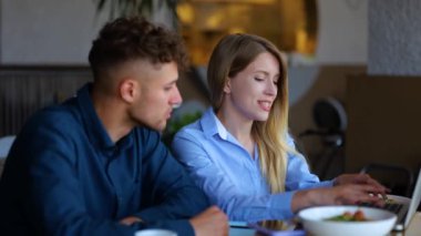 Close up of beautiful happy young woman sitting at table in restaurant with handsome man talking and typing on laptop. Caucasian male and female in cafeteria using computer and speaking. Communication