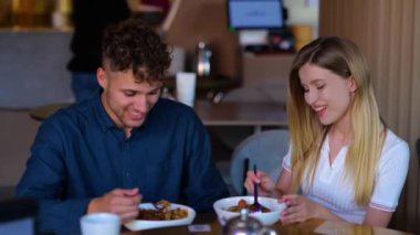 Close up of happy young girlfriend smiling and feeding her boyfriend while sitting in restaurant. Slow motion. Caucasian cheerful couple eating indoor on romantic date. Love relations. Dating concept