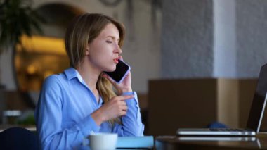 Caucasian beautiful young female speaking on smartphone and using laptop while sitting in restaurant. Close up of happy pretty businesswoman chatting on mobile phone. Slow motion. Side view