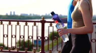 Close up of young athletic people in sportswear walking outdoors holding bottle of water in hands. Sporty athletes walk outside on bridge. Healthy activity. Couple after workout. Sport concept