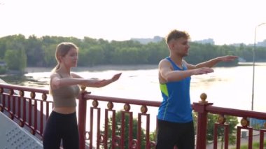 Portrait of young Caucasian couple exercising outdoor on the bridge with beautiful view on river. Happy man and woman stretching body and warming up muscles outside in city. Sport lifestyle concept