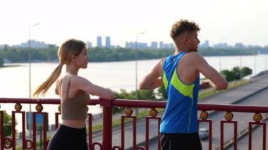 Portrait of young Caucasian couple man and woman exercising outdoor on the bridge turning body from side to side. Happy male and female stretching body and warming up muscles. Sport concept