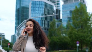 Close up of happy young businesswoman walking in street and speaking on mobile phone in city. Beautiful Caucasian female professional chatting on cellphone outdoors. Urban lifestyle. Business concept