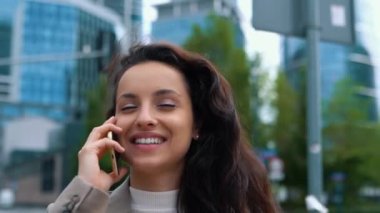 Close up of Caucasian young cheerful female walks in town while calling on smartphone. Joyful positive woman in city speaking on cellphone and smiling in good mood. Business style. Urban concept