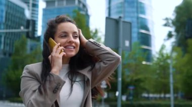 Camera moving around young Caucasian professional woman speaking on mobile phone. Happy beautiful positive female standing in street calling on smartphone. Business lifestyle. Close up