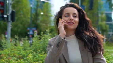 Close up of Caucasian young pretty female in suit sitting on a bench talking on cellphone outside in city. Beautiful professional woman having conversation on smartphone outdoor. Businesswoman concept