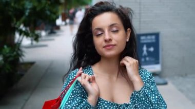 Close up of young Caucasian brunette woman walking outdoors with bags after shopping in good mood and touching her hair. Beautiful female buyer with purchases and packets on street. Buying concept