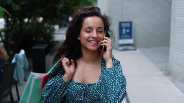 Close up portrait of young joyful woman speaking on cellphone and smiling while walking outdoors in city. Positive happy female walks in street with paper bags after buying stuff. Shopping concept