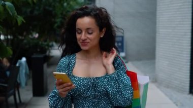 Close up shot of young happy beautiful woman texting on cellphone and smiling while walking outdoors in city. Positive Caucasian female walks in street with paper bags in good mood. Shopping concept