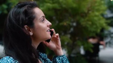 Side view of young attractive female smiling and speaking on mobile phone while walking in street. Close up of beautiful positive woman calling on cellphone outside in good mood. Communication concept