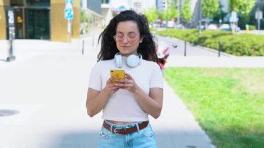 Pembe gözlüklü, akıllı telefonuyla mesajlaşan güneşli bir günde şehirde yürürken internette gezinen mutlu genç bir kadın. Sıradan, pozitif bir kadının cep telefonlu portresi. Yavaş çekim